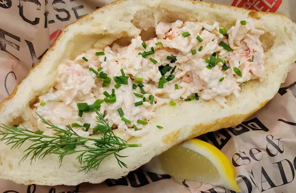 Lobster roll sandwich with chopped lobster salad garnished with chopped chives, served in a toasted bun with a lemon wedge and dill on a sheet of parchment paper.